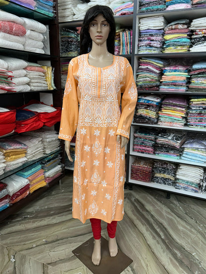 An orange long kurta with white floral embroidery displayed on a mannequin in a clothing store. The kurta features a round neckline, three-quarter sleeves, and white embroidered patterns on the front and cuffs.