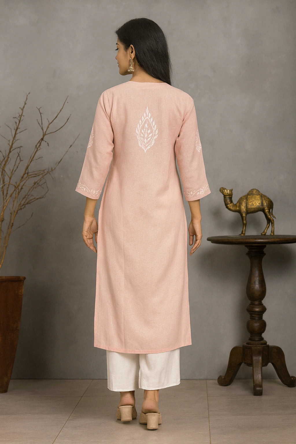 Woman wearing a pink kurta with white embroidery, standing in a room with a wooden table and decorative camel statue.
