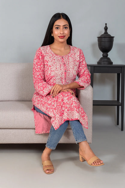 Lucknowi chikankari A woman sits on a light gray sofa wearing a pink and white floral printed kurta with blue jeans and beige heeled sandals. A dark side table with a decorative urn is visible in the background against a light gray wall.