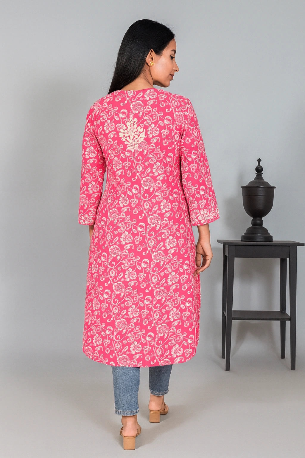 Lucknowi chiknkari A woman shown from the back wearing a pink long kurta with a white floral block print pattern, paired with blue jeans and beige heels, standing in a studio next to a black side table.