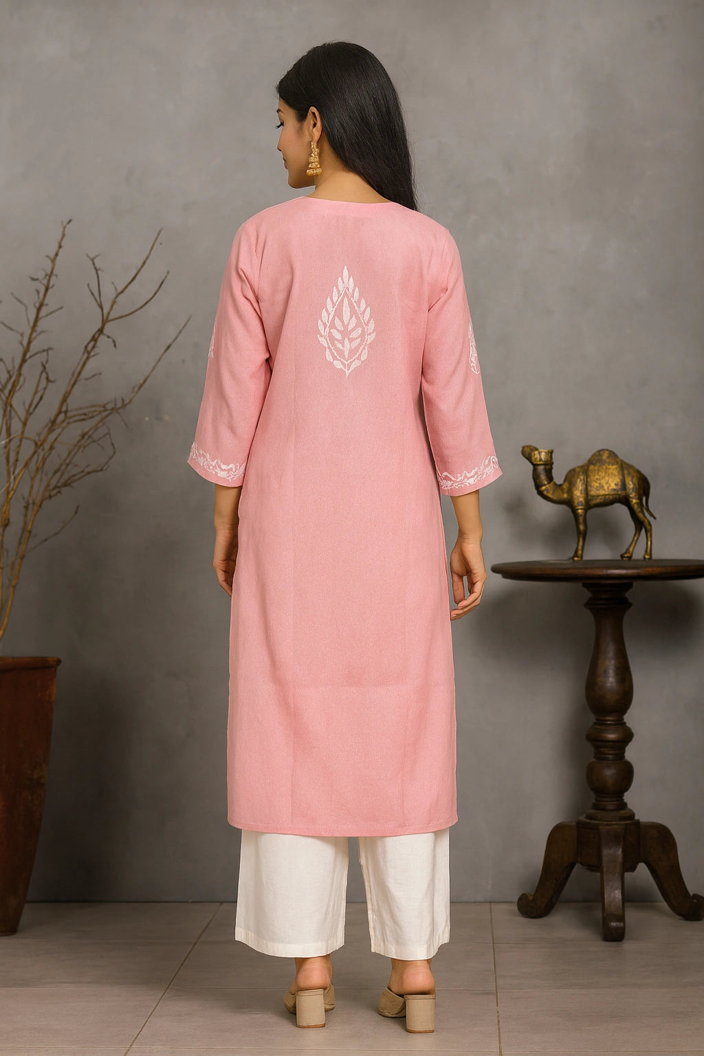 Woman wearing a pink kurta with white embroidery, standing in a room with a gray wall and wooden table.