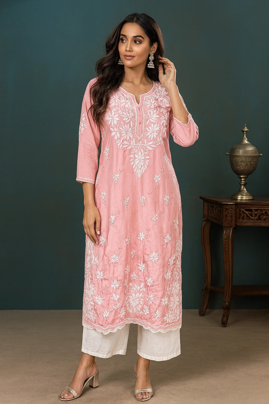 Woman wearing a pink embroidered kurta with white pants against a dark green wall.