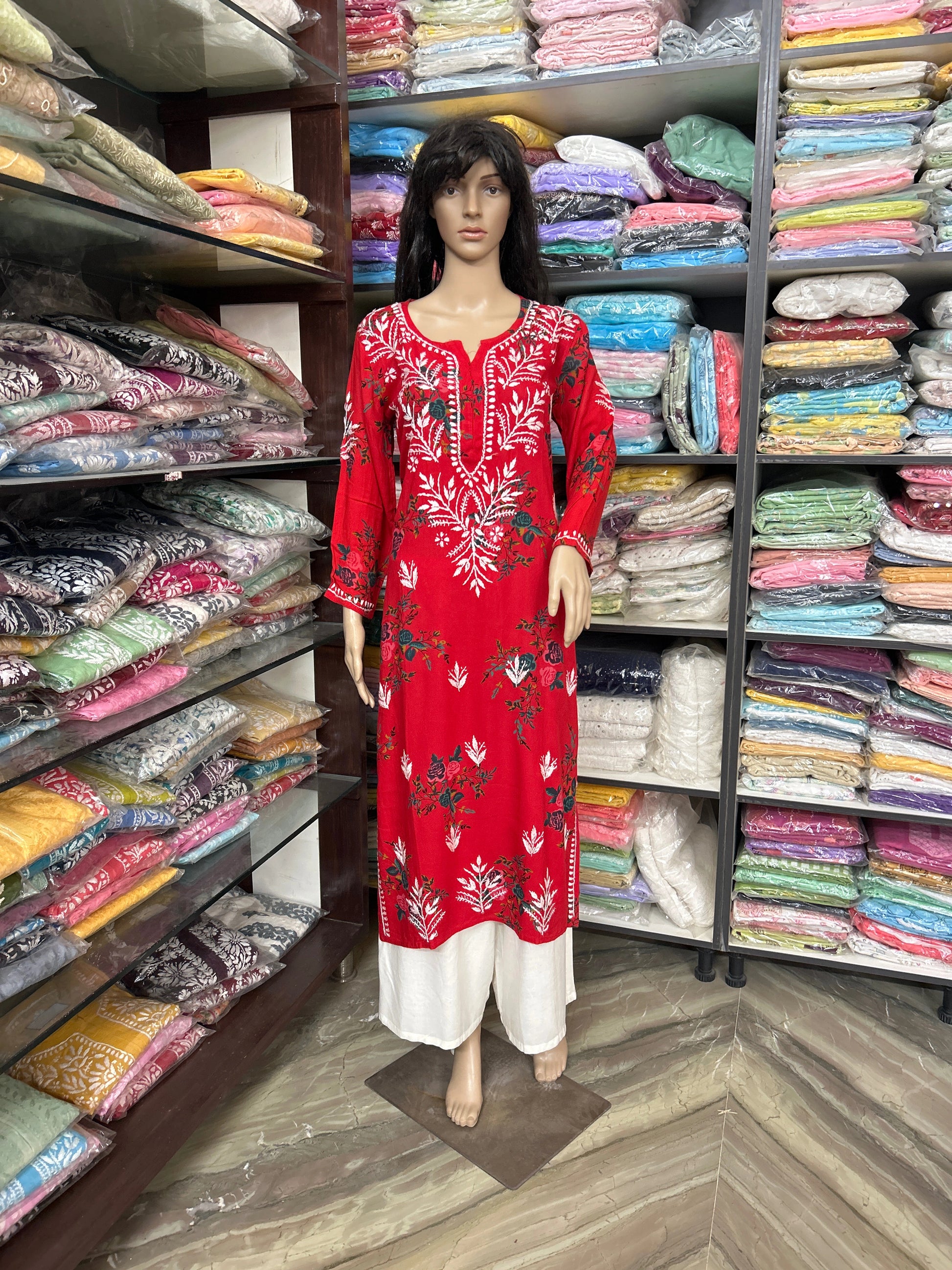 Mannequin wearing a red embroidered dress in a fabric store with shelves of colorful fabrics.