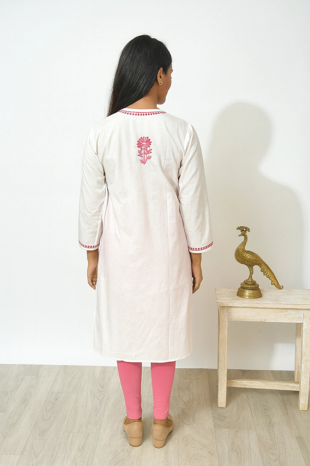 Woman wearing a white kurta with pink floral embroidery, standing in a minimalistic room.