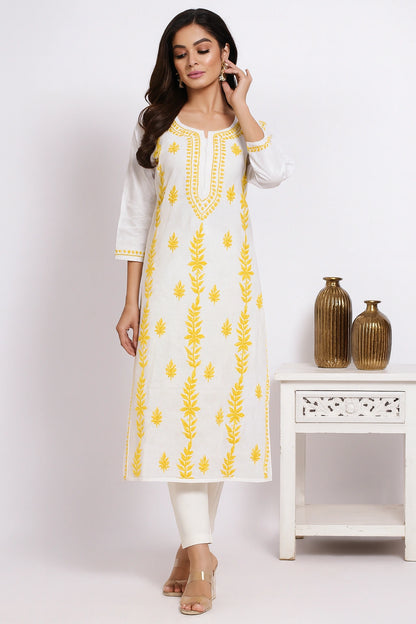 Woman wearing a white kurta with yellow embroidery in a minimalistic setting.