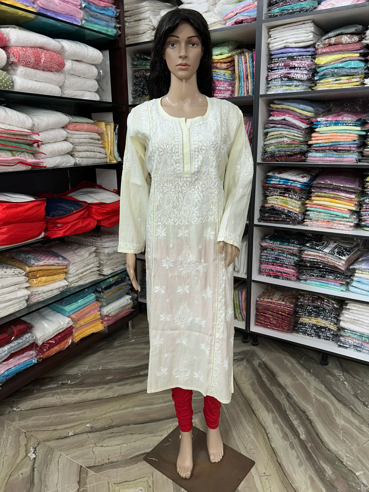A cream-colored long kurta with white floral embroidery, displayed on a female mannequin standing in a clothing store. The mannequin is also wearing red leggings, and shelves of folded garments are visible in the background.