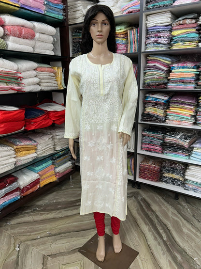 A cream-colored long kurta with white floral embroidery, displayed on a female mannequin standing in a clothing store. The mannequin is also wearing red leggings, and shelves of folded garments are visible in the background.