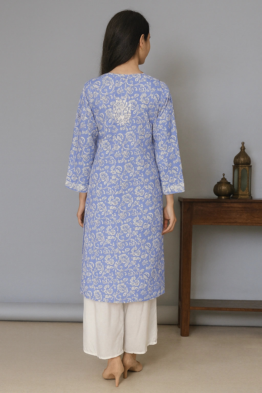 Lucknowi chikankari Woman wearing a blue floral kurta with white pants standing in a room with a wooden table and decorative items.