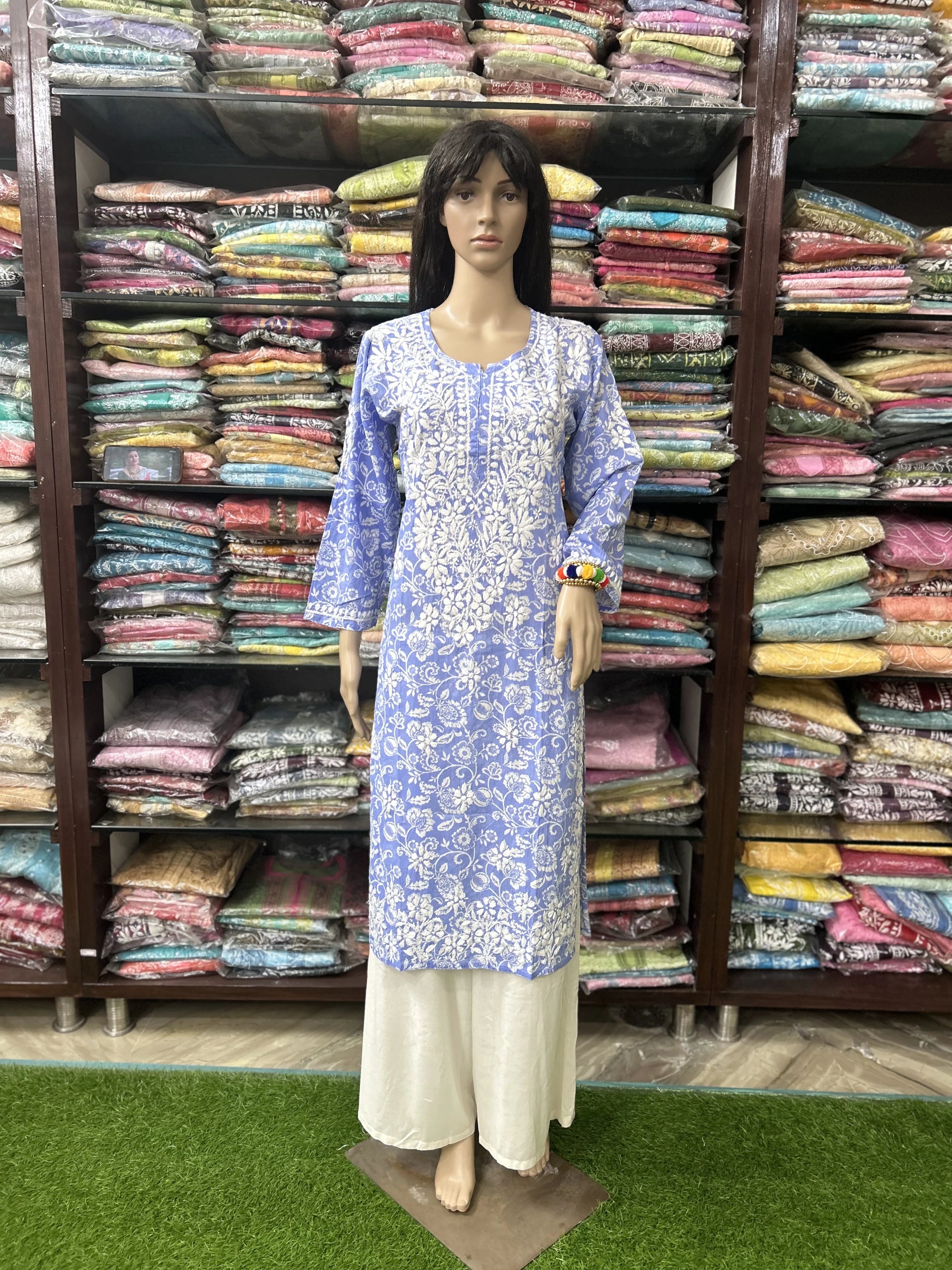Lucknowi Chikankari Mannequin wearing a blue and white patterned dress in front of shelves filled with colorful fabrics.