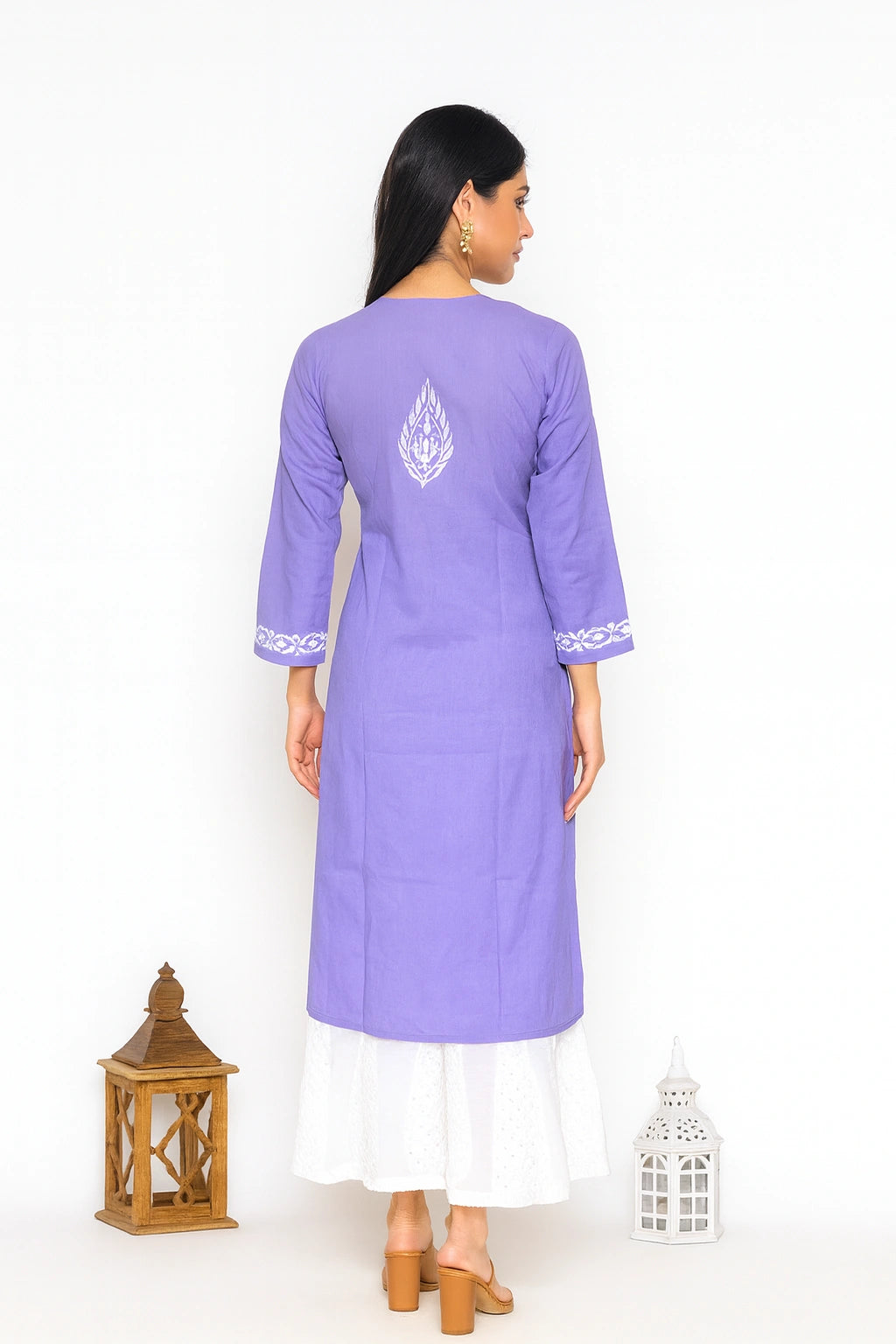 A woman stands with her back to the camera wearing a lavender purple kurta with white embroidery on the back and cuffs, layered over a white skirt. She is photographed against a plain white studio background with decorative lantern props on the floor.