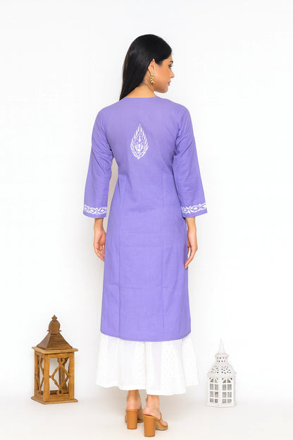 A woman stands with her back to the camera wearing a lavender purple kurta with white embroidery on the back and cuffs, layered over a white skirt. She is photographed against a plain white studio background with decorative lantern props on the floor.
