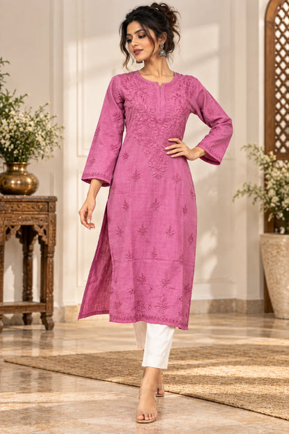 A woman models a three-quarter sleeve magenta pink embroidered kurta with white pants in an indoor setting with a tiled floor and decorative plants.