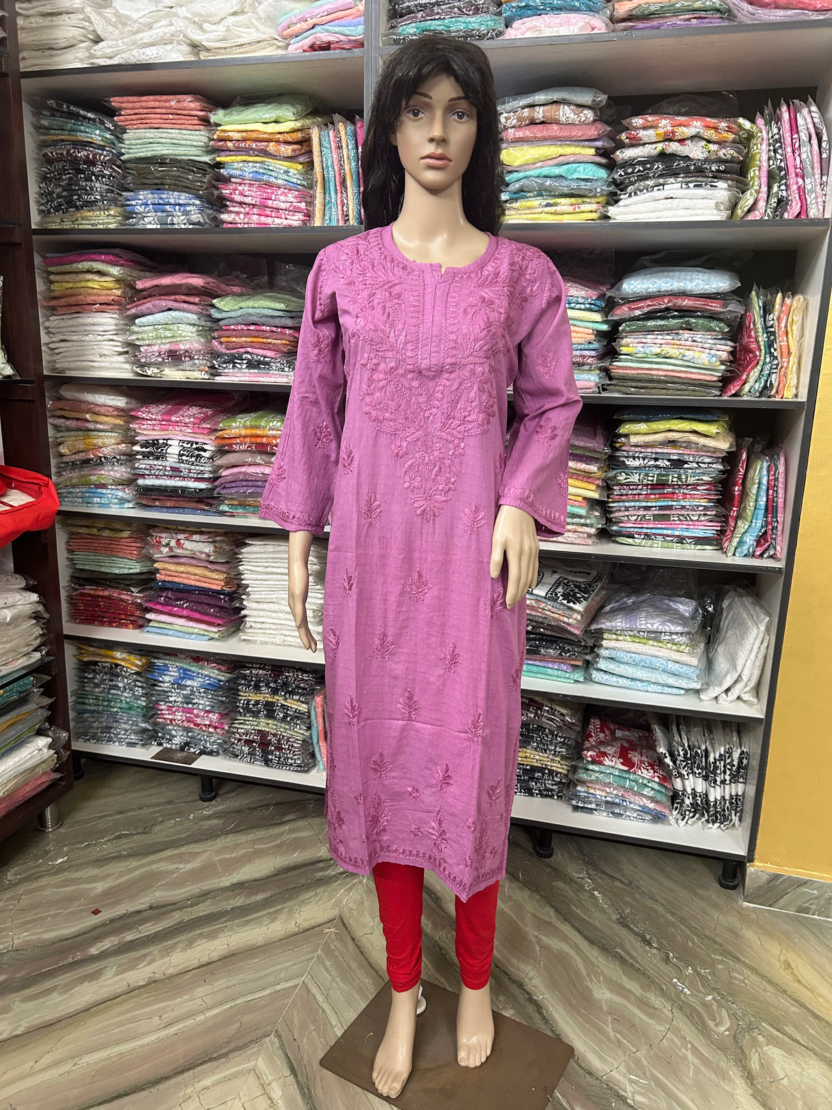 A mannequin wearing a pink embroidered kurta with red leggings, standing in front of shelves filled with folded colorful fabrics in a retail store.