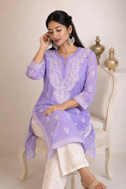 A woman seated on a white upholstered chair wearing a lavender embroidered kurta with white pants, against a light beige studio background with decorative gold vases.