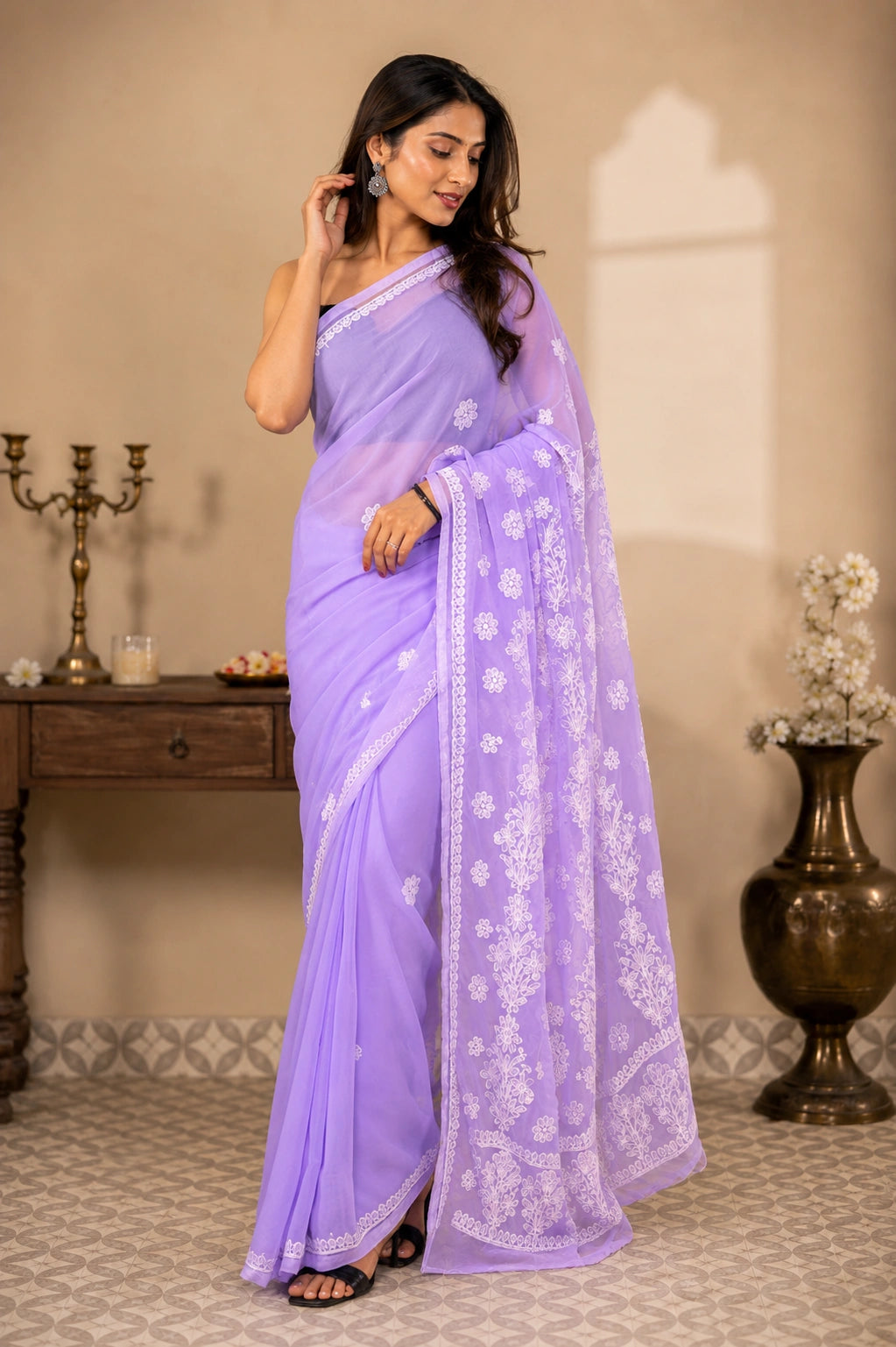 A woman models a lavender purple saree with white floral embroidery, standing in a decorated indoor setting with a wooden table and decorative vase in the background.