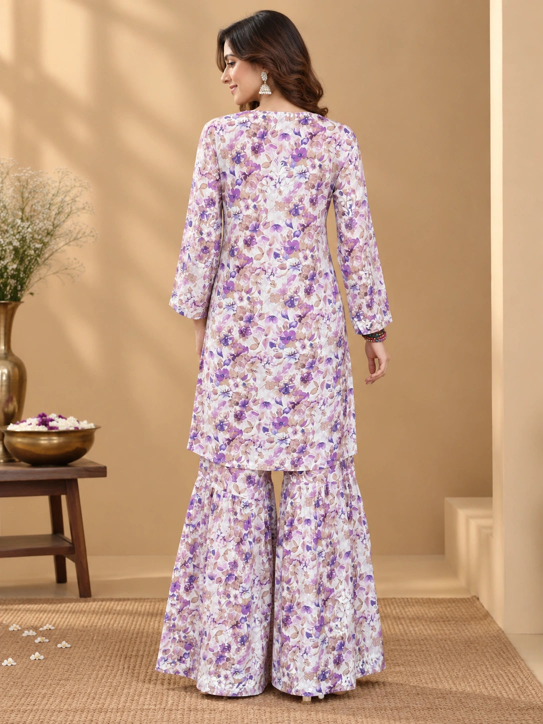 Lucknowi chikankari A woman stands with her back to the camera wearing a floral printed kurta and flared pants set in a warm beige indoor setting with decorative plants and a wooden table.