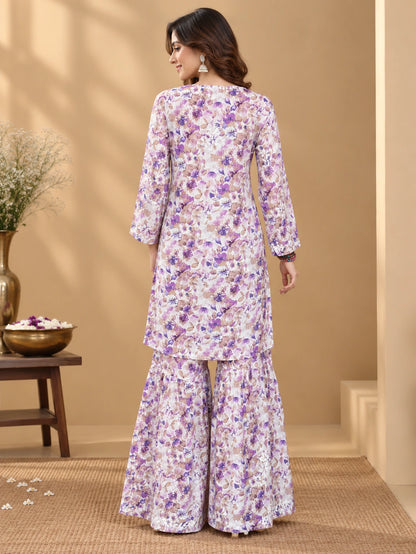 Lucknowi chikankari A woman stands with her back to the camera wearing a floral printed kurta and flared pants set in a warm beige indoor setting with decorative plants and a wooden table.
