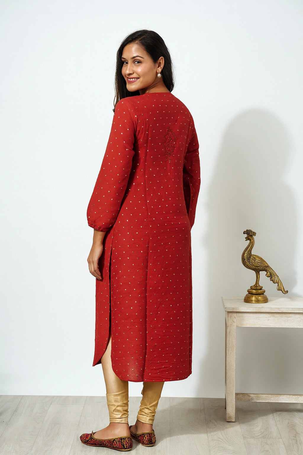 lucknowi chikankari A woman modeling a red long kurta with small gold polka dots, paired with beige leggings and brown patterned flat shoes, standing in a studio setting next to a small table with a decorative peacock statue.