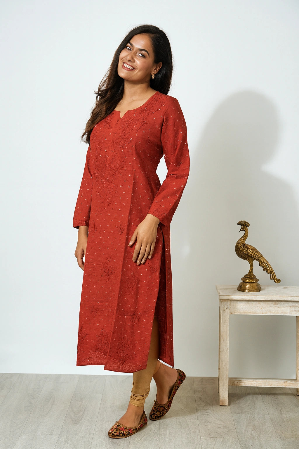 lucknowi chikankari A woman modeling a rust-red long-sleeve kurta dress with a subtle dotted pattern, standing in a studio setting next to a small table with a decorative peacock figurine.