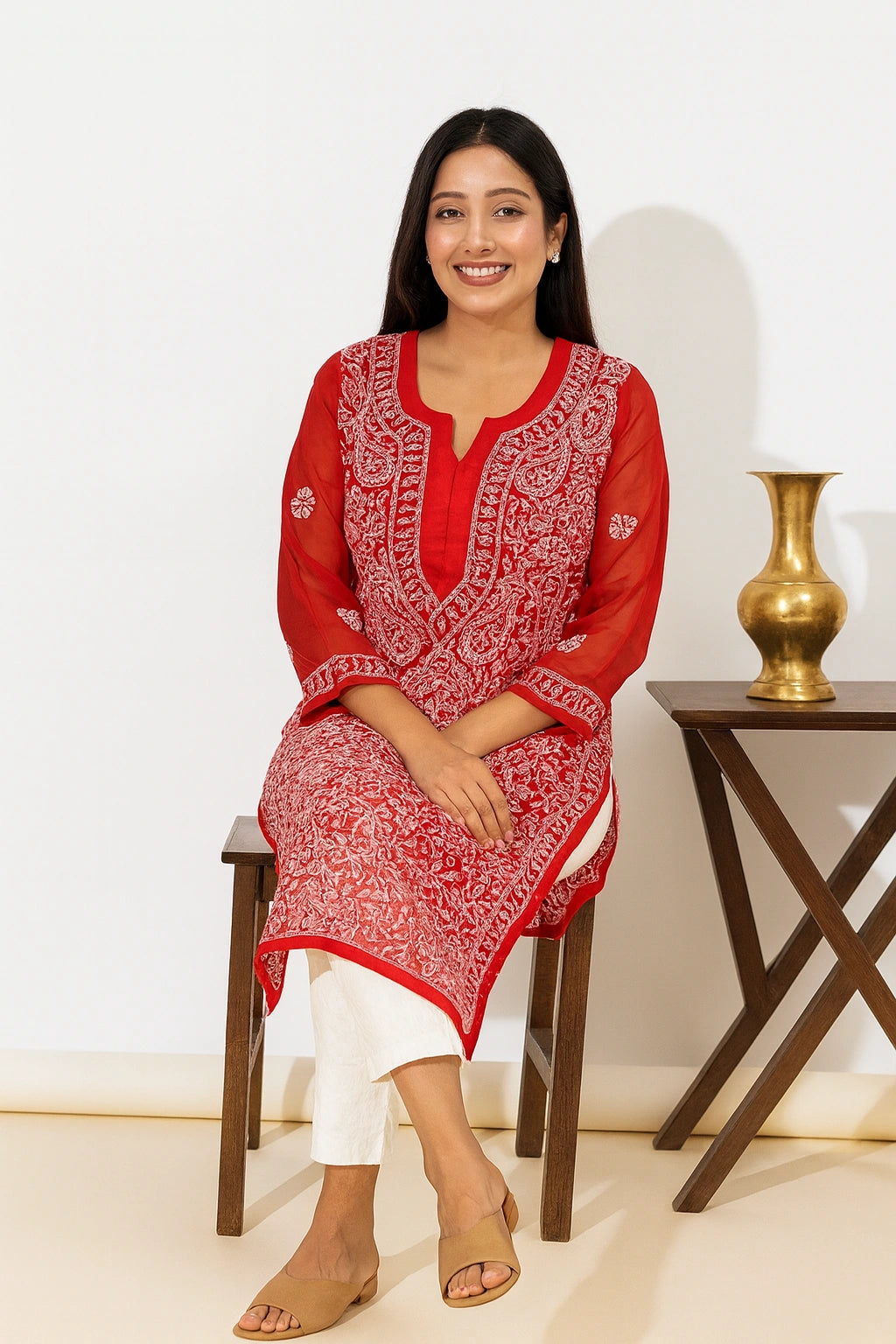 A woman sits on a wooden stool wearing a red kurta with intricate white embroidery, paired with white pants and beige sandals, in a brightly lit indoor setting.