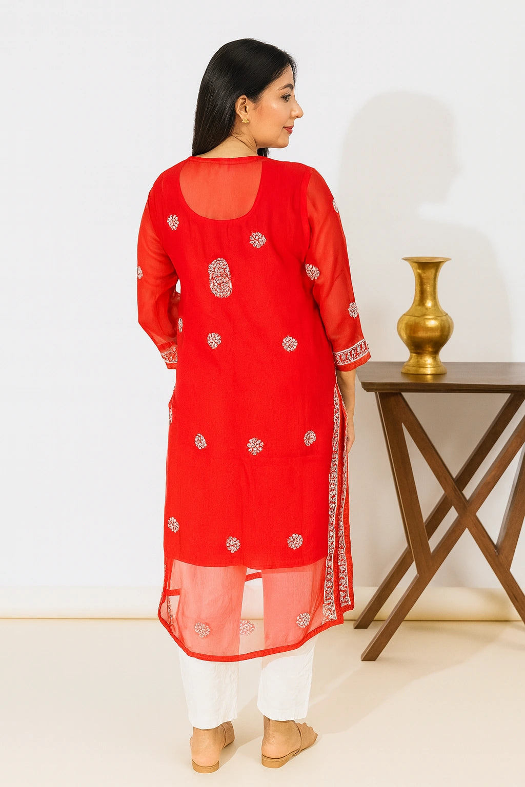 A woman is shown from the back wearing a bright red traditional kurta with white pants and beige flat shoes. The kurta features white floral embroidery and a sheer mesh overlay.