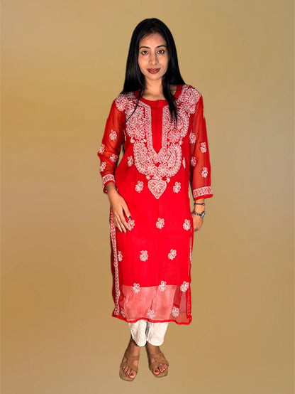 A woman modeling a red long kurta with intricate white embroidery, paired with white pants and brown sandals, set against a solid beige background.