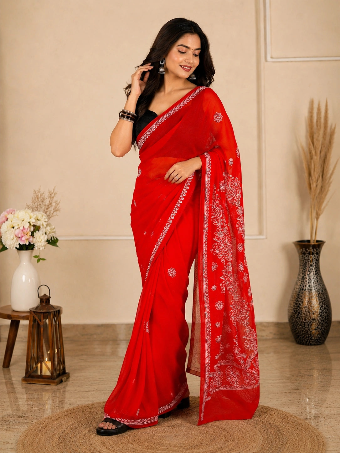 A woman modeling a bright red saree with white floral embroidery and a black blouse, standing in a styled indoor setting with decorative vases and a lantern.