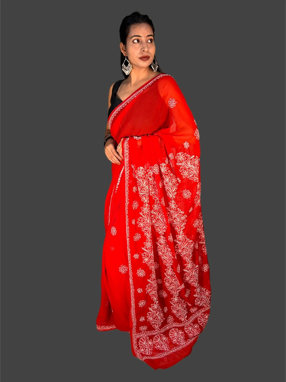 A woman modeling a bright red saree with intricate white floral embroidery and a black sleeveless blouse, set against a solid dark gray background.