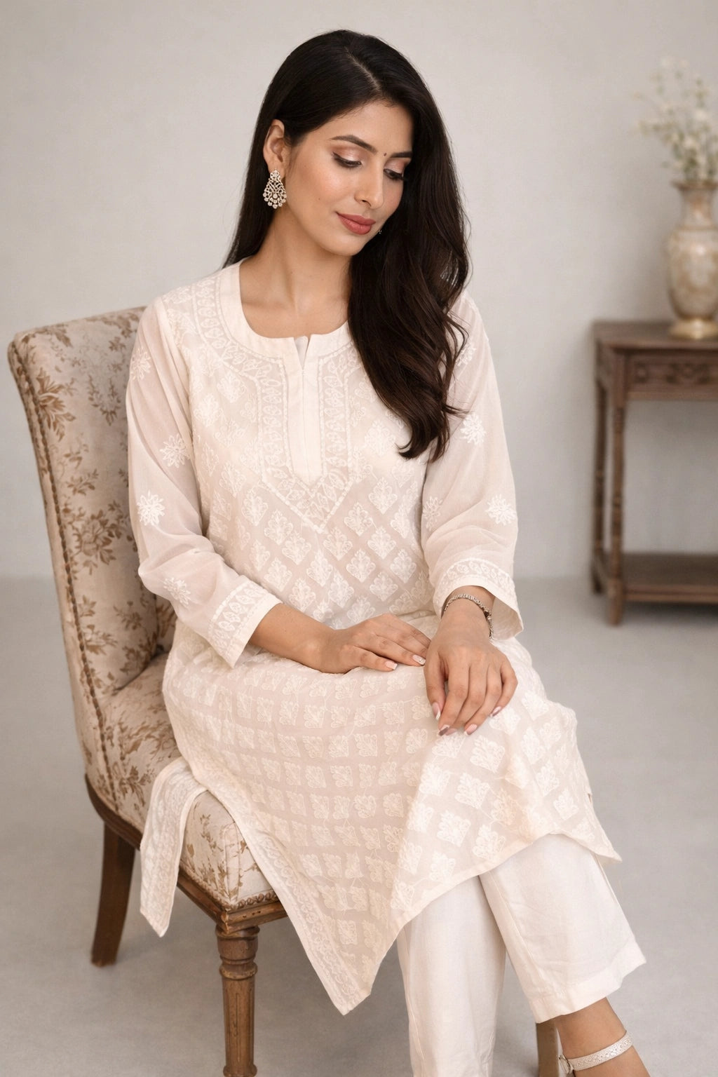 A woman seated in an upholstered armchair wearing a light cream or off-white embroidered kurta with matching pants. The setting is a softly lit indoor room with a side table and vase in the background.
