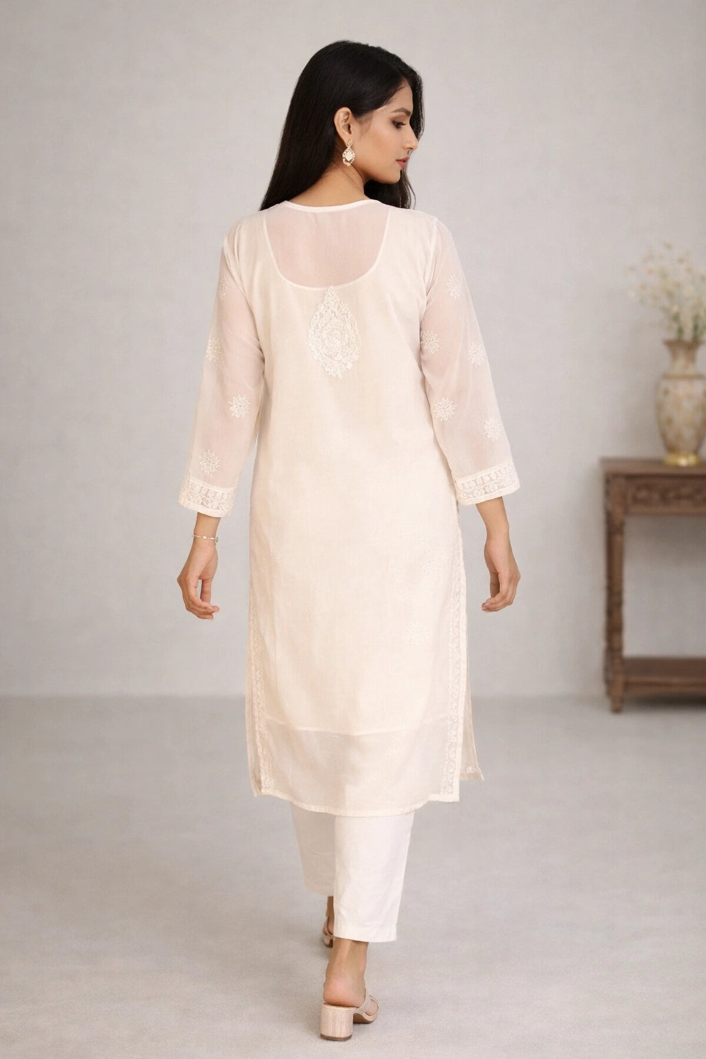 Lucknowi chikankari A woman wearing a light cream/off-white kurta with white pants, shown from the back in a studio setting with a neutral background and a side table.
