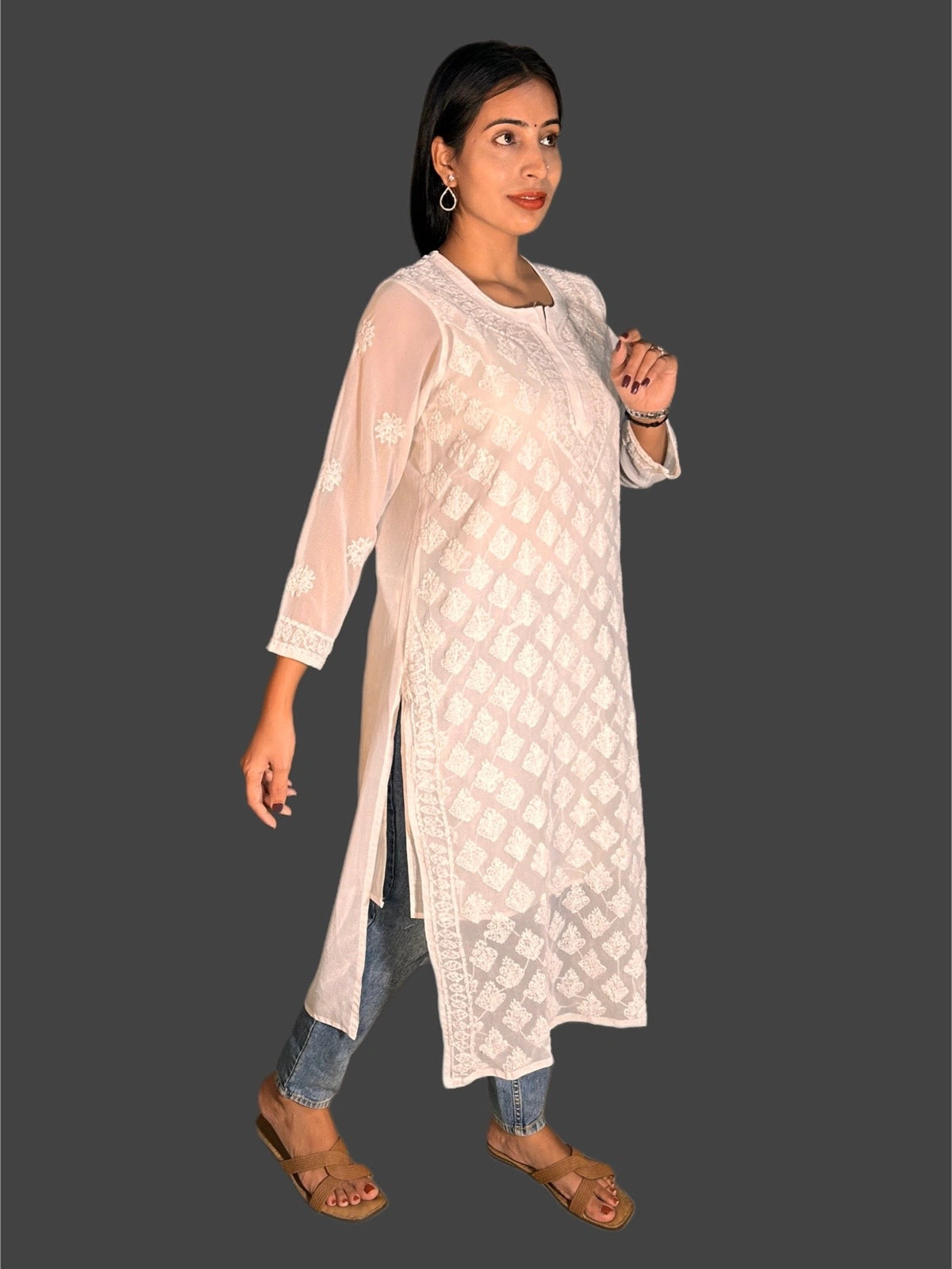 A woman modeling a white embroidered kurta with sheer sleeves and blue jeans, standing against a solid dark gray background.