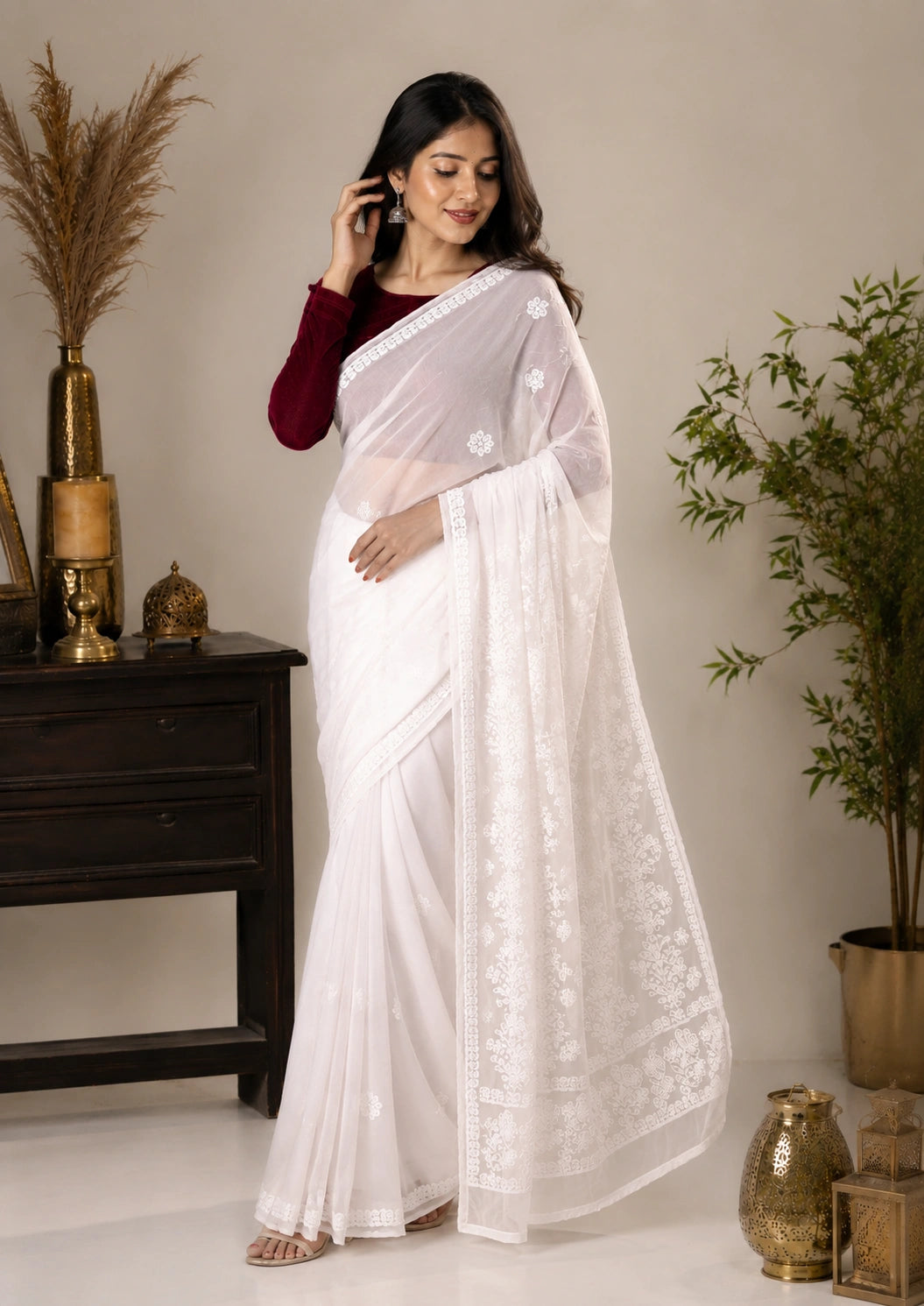 A woman models a white saree with intricate floral embroidery and a deep maroon long-sleeve blouse, standing in a styled indoor setting with plants and decorative vases.