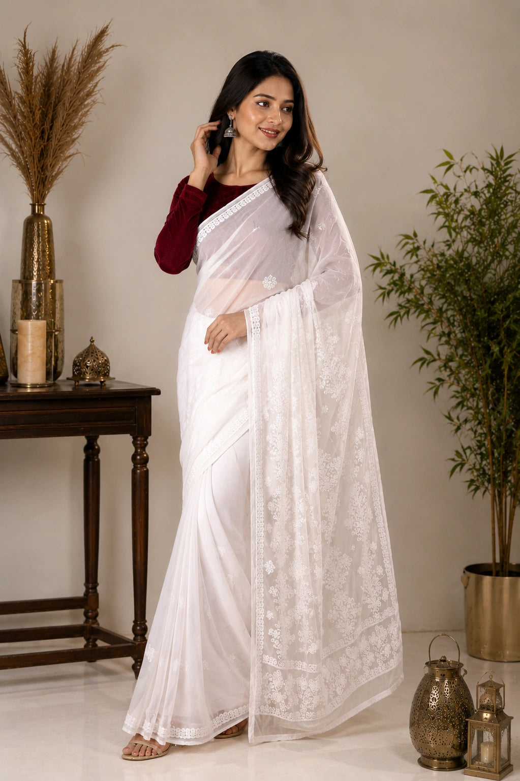 A woman models a white saree with intricate floral embroidery along the border and pallu, paired with a dark red long-sleeve blouse. She is standing in a styled indoor setting with decorative plants and a wooden table.