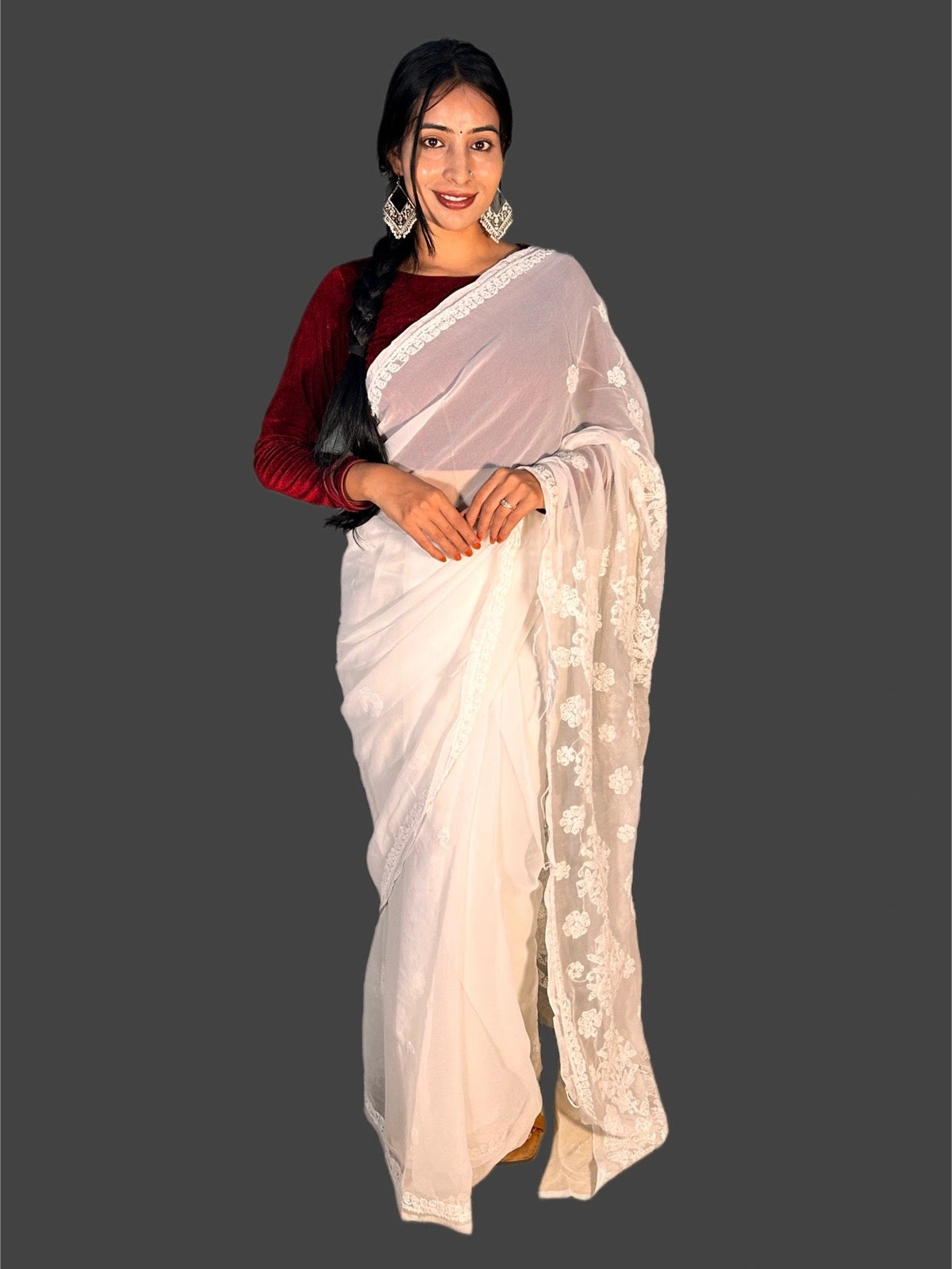 A woman modeling a white saree with floral embroidery on the border, paired with a dark red long-sleeve blouse, set against a solid dark gray background.