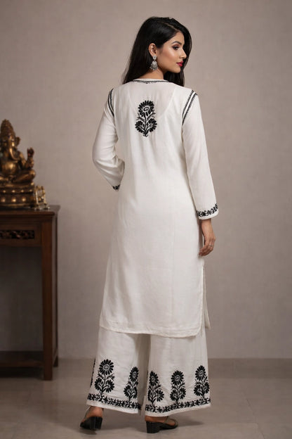 Woman wearing a white outfit with black floral embroidery against a neutral background