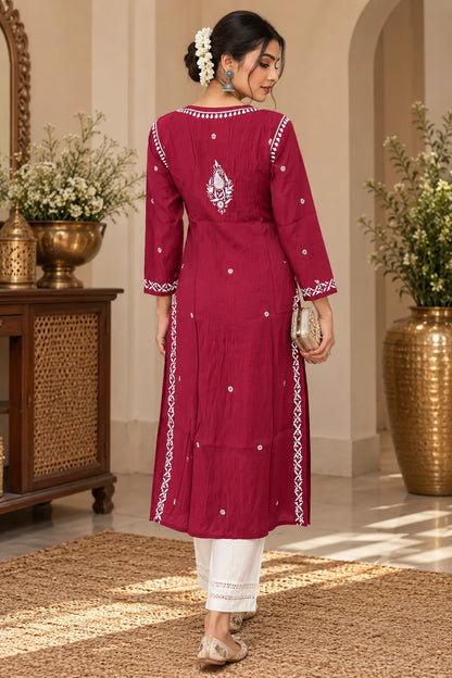 A woman wearing a wine-colored embroidered kurta with white pants, standing in front of a carved wooden door with floral decorations.