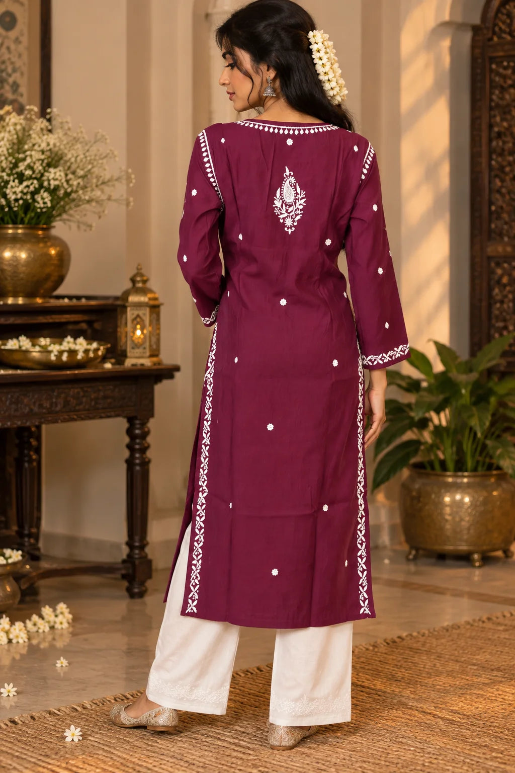 A woman wearing a wine-colored embroidered kurta with white pants, standing in front of a carved wooden door with floral decorations.