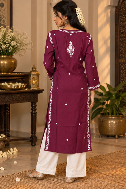 A woman wearing a wine-colored embroidered kurta with white pants, standing in front of a carved wooden door with floral decorations.