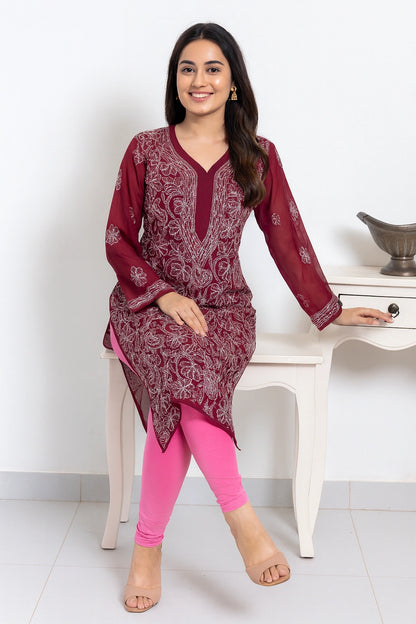 A woman models a three-quarter sleeve maroon kurta with white floral embroidery, paired with bright pink leggings, sitting on a white wooden chair in a clean indoor setting.