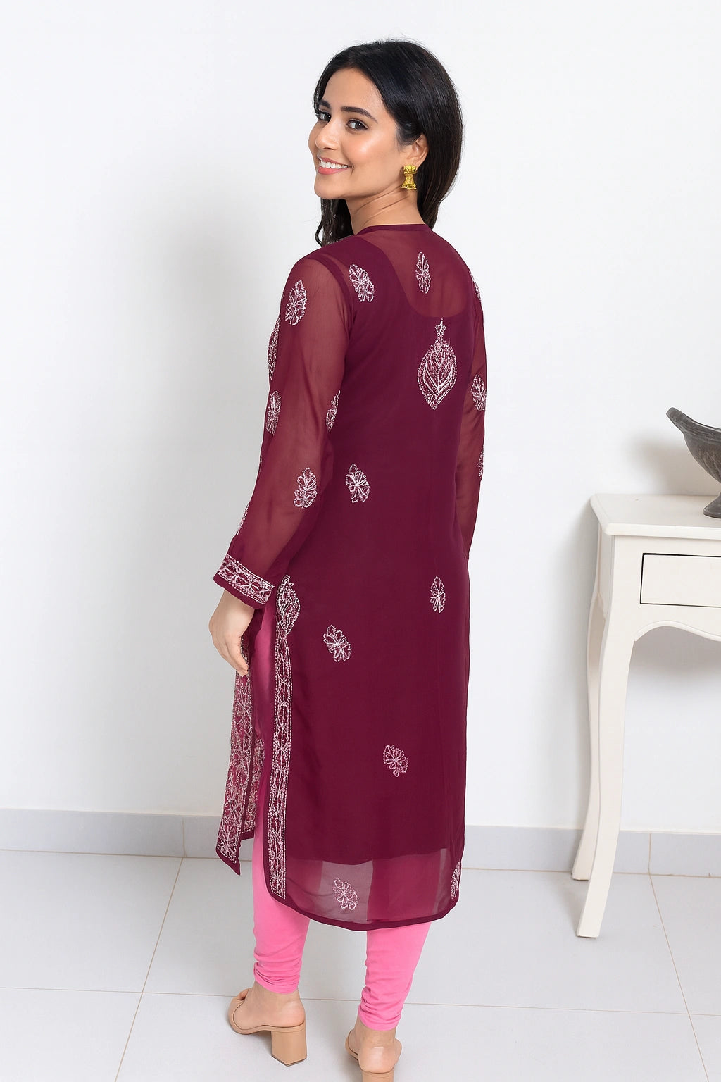 Lucknowi chikankari A woman modeling a deep maroon sheer georgette kurta with white printed motifs, paired with pink leggings and beige heels, shown from a three-quarter back view in an indoor setting.