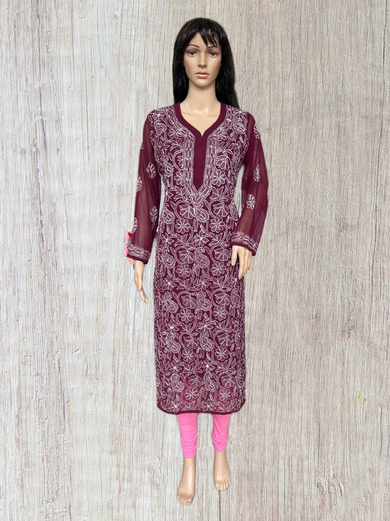 A mannequin wearing a long-sleeved maroon embroidered kurta with white floral patterns, paired with bright pink leggings, set against a light wood-textured background.