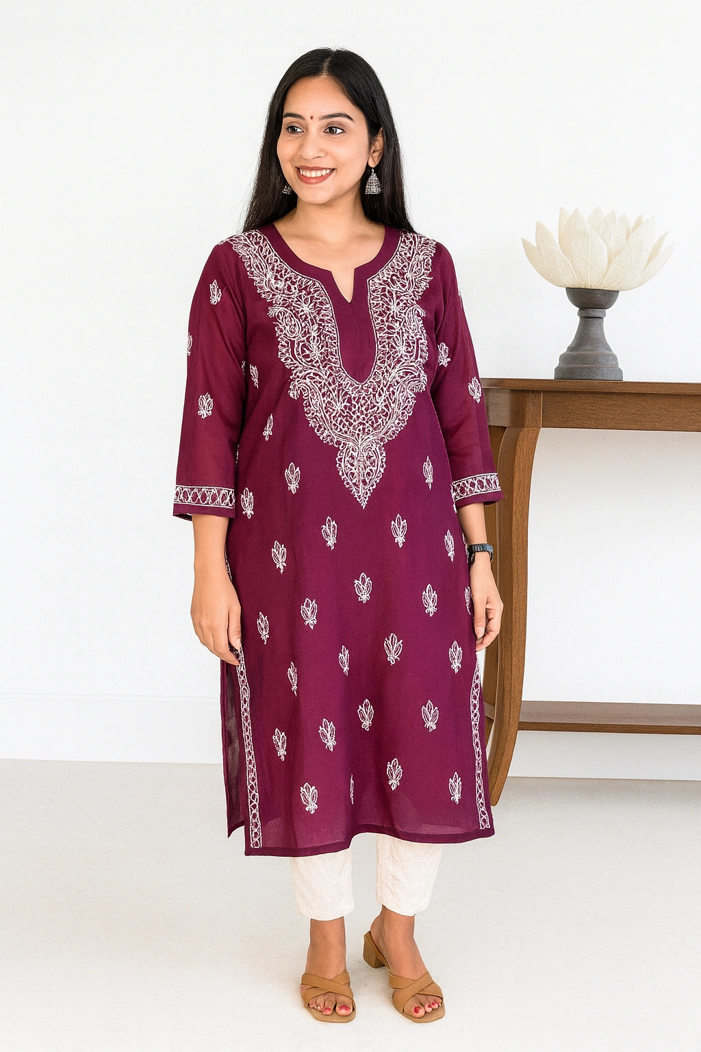 A woman modeling a deep maroon kurta with white embroidery and white printed motifs, paired with white pants and brown sandals, standing in a bright studio setting.
