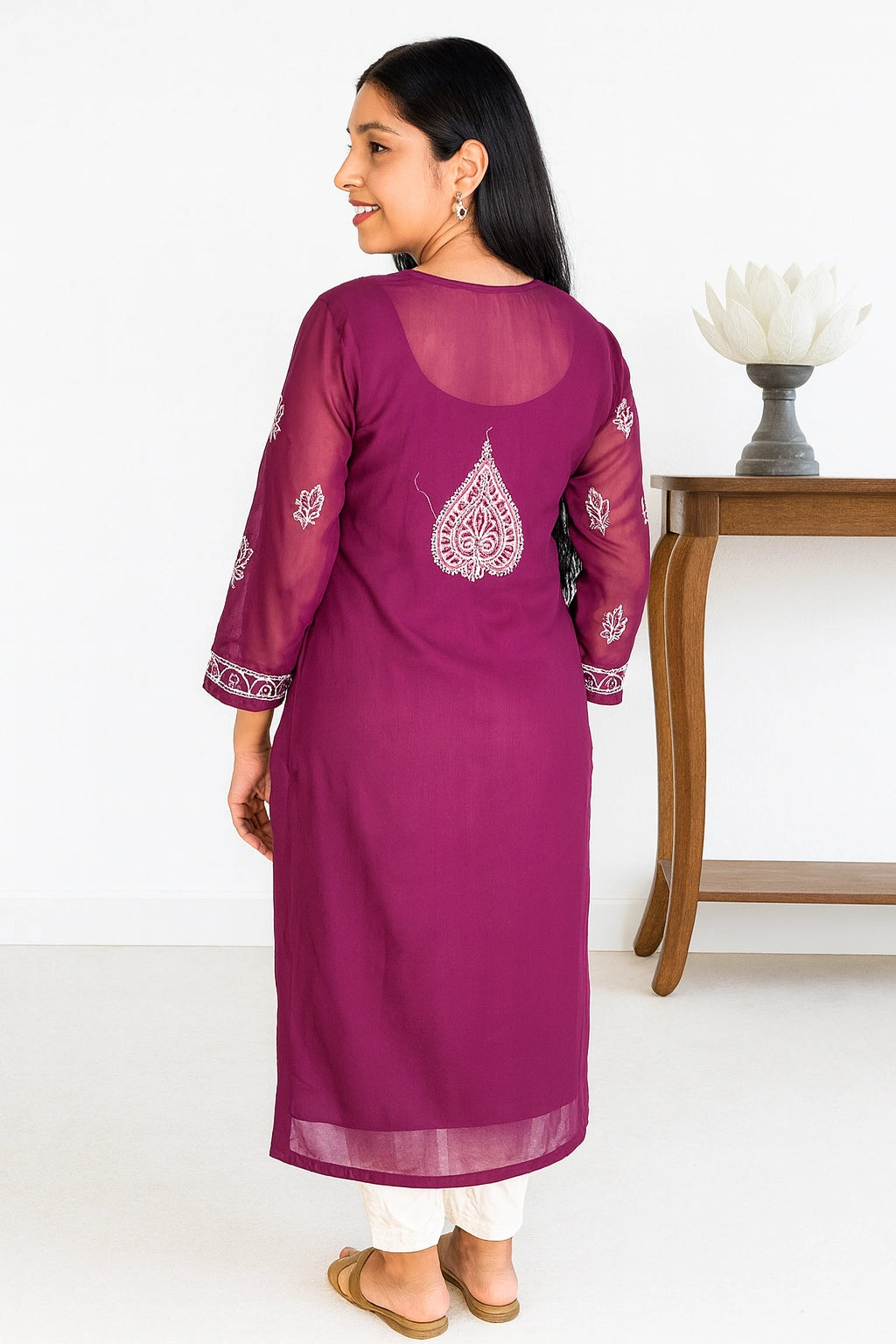 A woman wearing a magenta purple kurta with white embroidery, shown from the back. She is standing in a bright room next to a wooden side table with a white lotus vase.