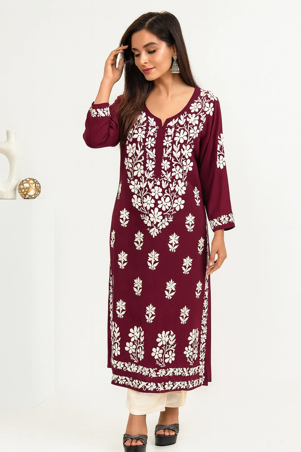 A woman modeling a deep maroon long kurta with intricate white floral embroidery, paired with cream pants and black sandals, set against a clean white studio background.