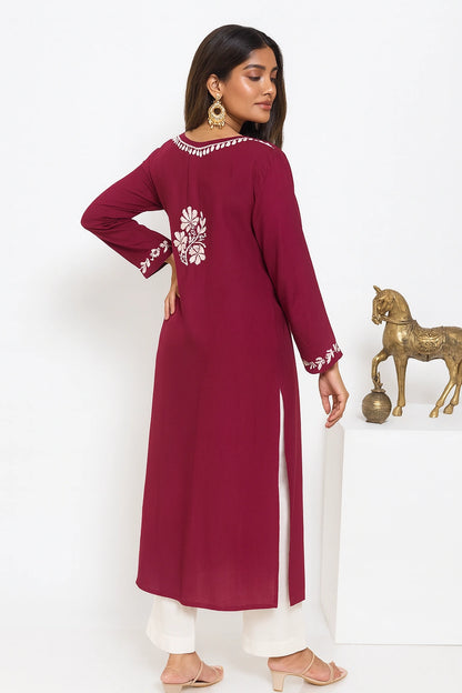 A woman modeling a long-sleeved maroon kurta with white floral embroidery on the back and sleeve cuffs, paired with white pants, in a studio setting with a gold horse statue.