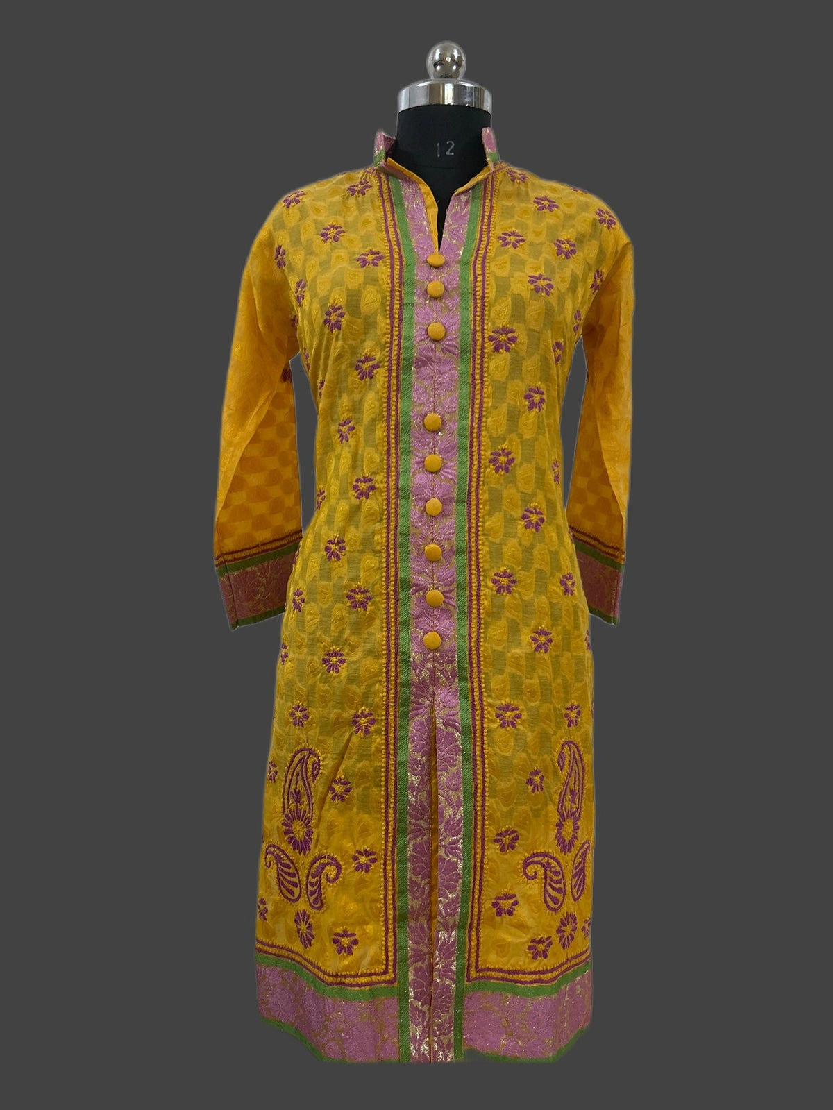 A yellow embroidered tunic with pink and green detailing, displayed on a black mannequin against a dark gray background.
