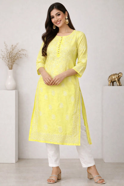 Lucknowi chikankari A woman modeling a light yellow kurta with white pants and silver sandals in a bright, modern studio setting with decorative white pedestals.