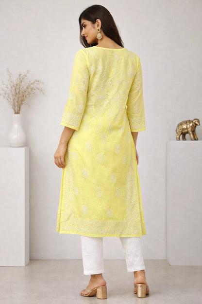 A woman wearing a light yellow embroidered kurta with white pants and beige block-heel sandals, shown from the back in a minimalist indoor setting.