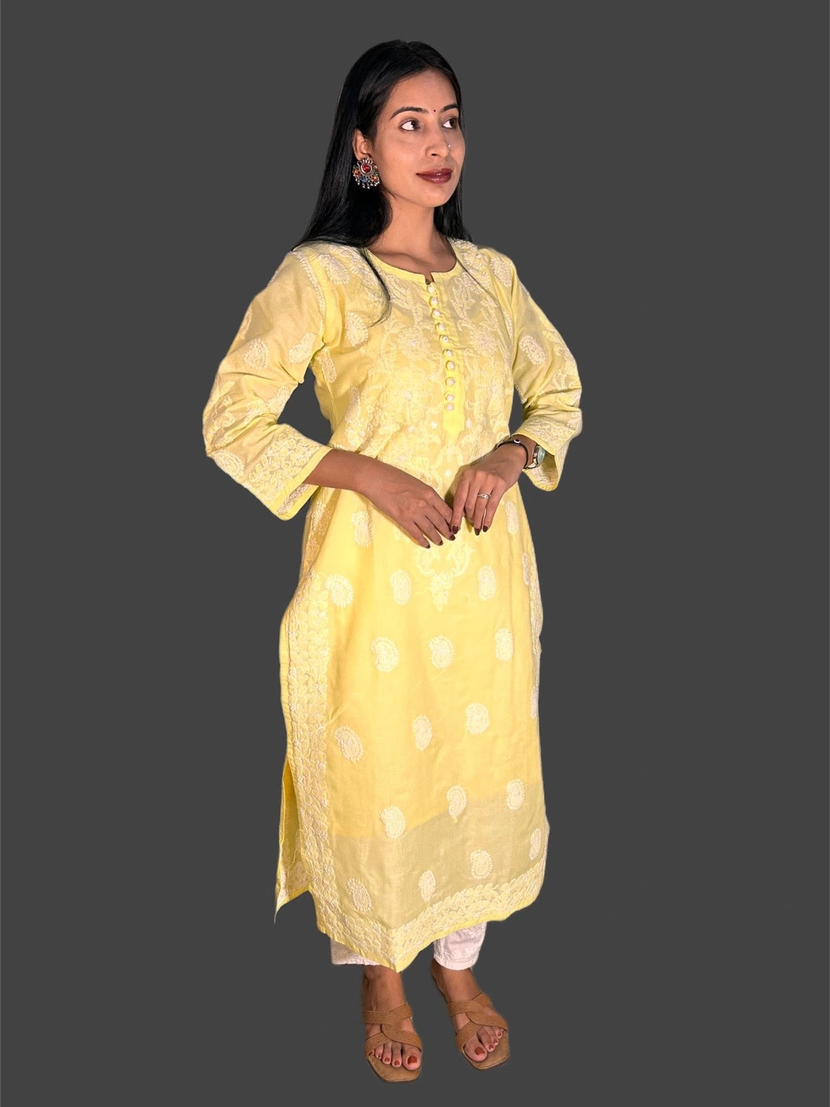 A woman modeling a light yellow kurta with white polka dots and white embroidered borders, paired with white pants, against a solid dark gray background.