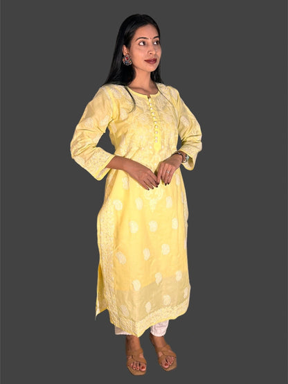 A woman modeling a light yellow kurta with white polka dots and white embroidered borders, paired with white pants, against a solid dark gray background.